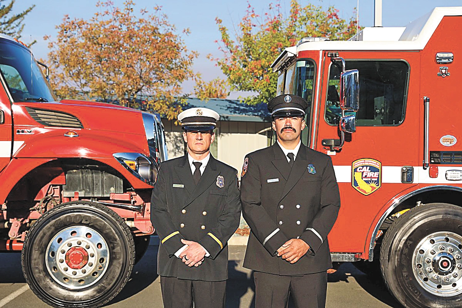 Two uniformed individuals stand with hands clasped between two red fire trucks, one marked "CAL FIRE", under autumn trees.