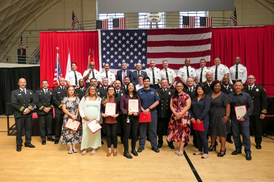Group of people in uniforms and civilian attire pose with certificates in gymnasium with red curtains and American flags.