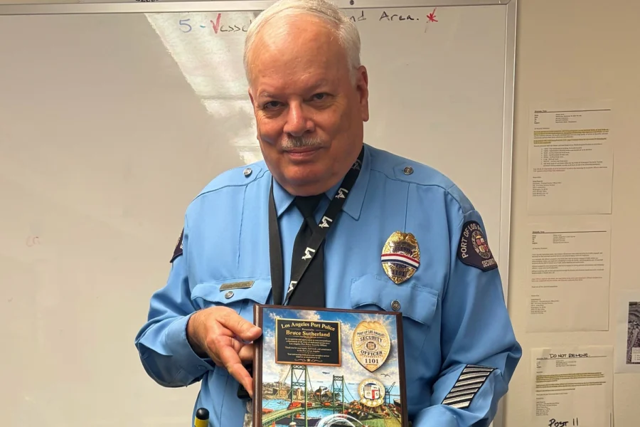 Person in blue police uniform holds framed plaque and glasses, standing in room with whiteboard, papers on wall, and table.