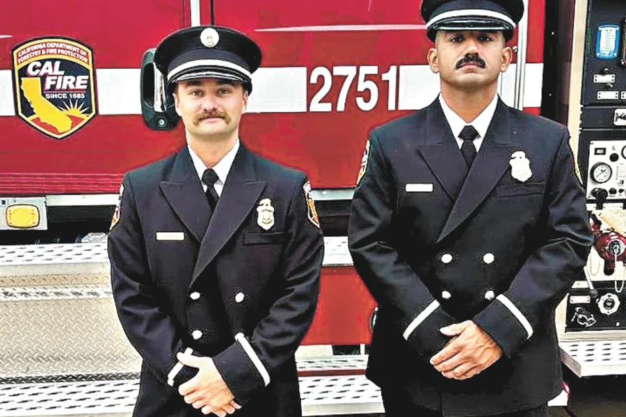 Two men in dark fire service uniforms stand with clasped hands in front of a red CAL FIRE fire truck marked 2751.
