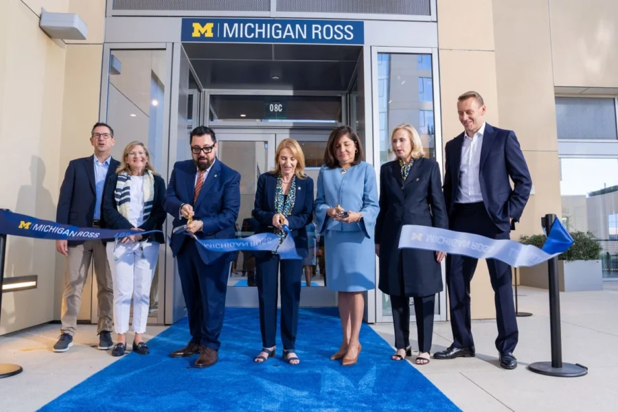 Six people stand on a blue carpet outside a building entrance, holding a blue ribbon with "MICHIGAN ROSS" text for a ribbon-cutting ceremony.