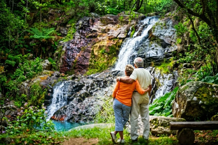 An older couple stands close, watching water cascade over rocks in a lush green forest.