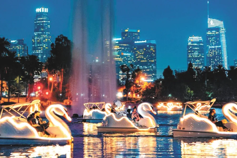 People ride illuminated swan boats on a lake at night, with a city skyline and fountain behind.