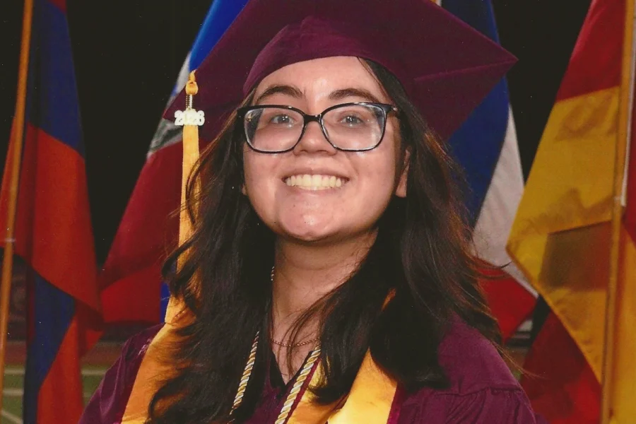 A person in maroon graduation cap and gown with yellow "ACOM" stole stands before flags, hill, and pink sky.