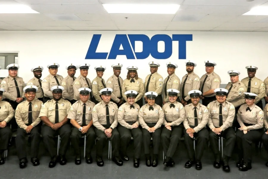 Group of uniformed personnel in beige shirts and white hats pose in front of a wall with "LADOT" signage, flags visible, office setting.