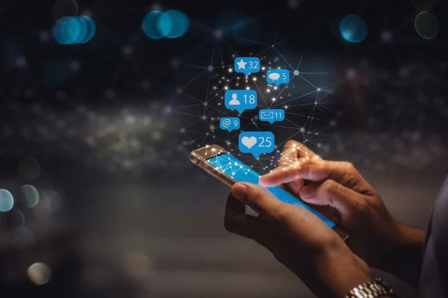 Hands hold a smartphone with glowing social media icons floating above it against a dark, blurred background.