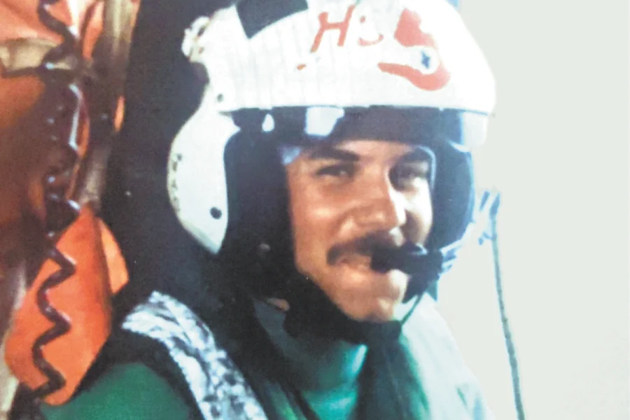 Person wearing white helmet with "H" and red heart, green shirt, vest; another in orange with coiled cord, indoor setting.
