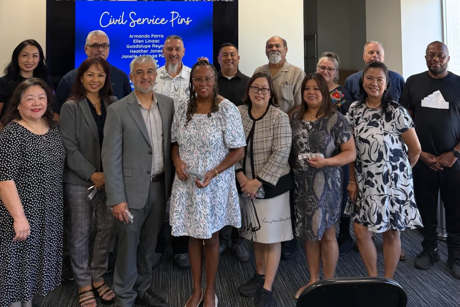 Group of people posing for a photo in an office with a screen showing "Civil Service Pins" and listed names.
