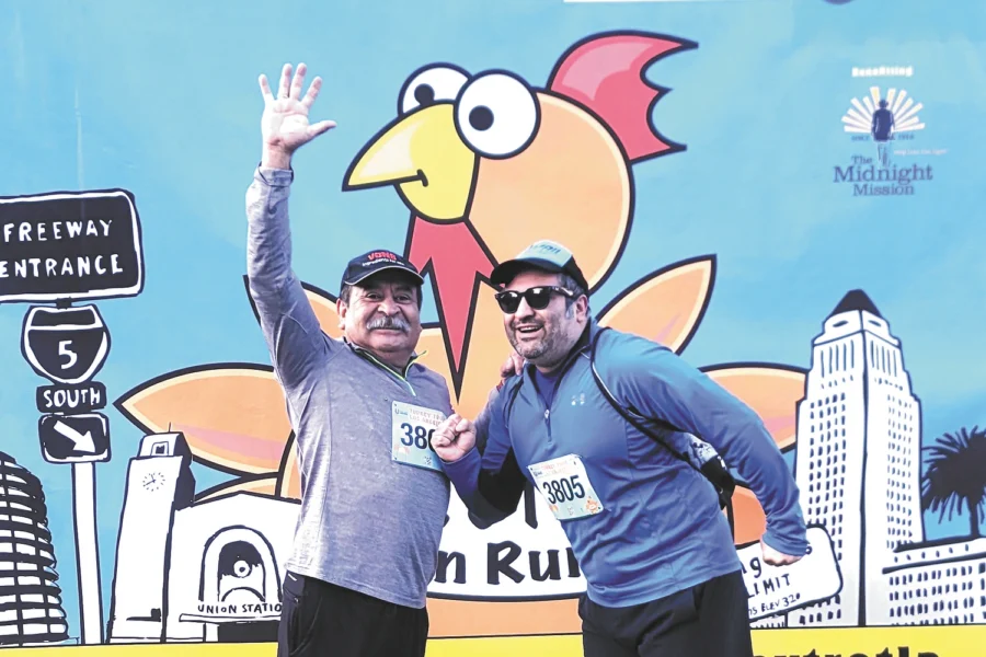 Two men wearing race bibs wave and pose on grass before a Turkey Trot Los Angeles banner with a cartoon turkey.