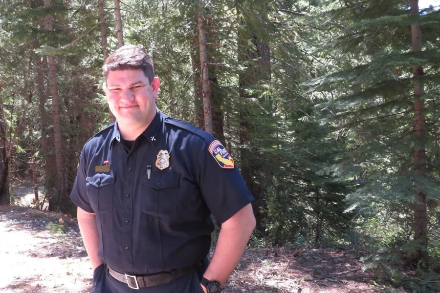 Man in uniform stands with hands in pockets among tall pine trees.