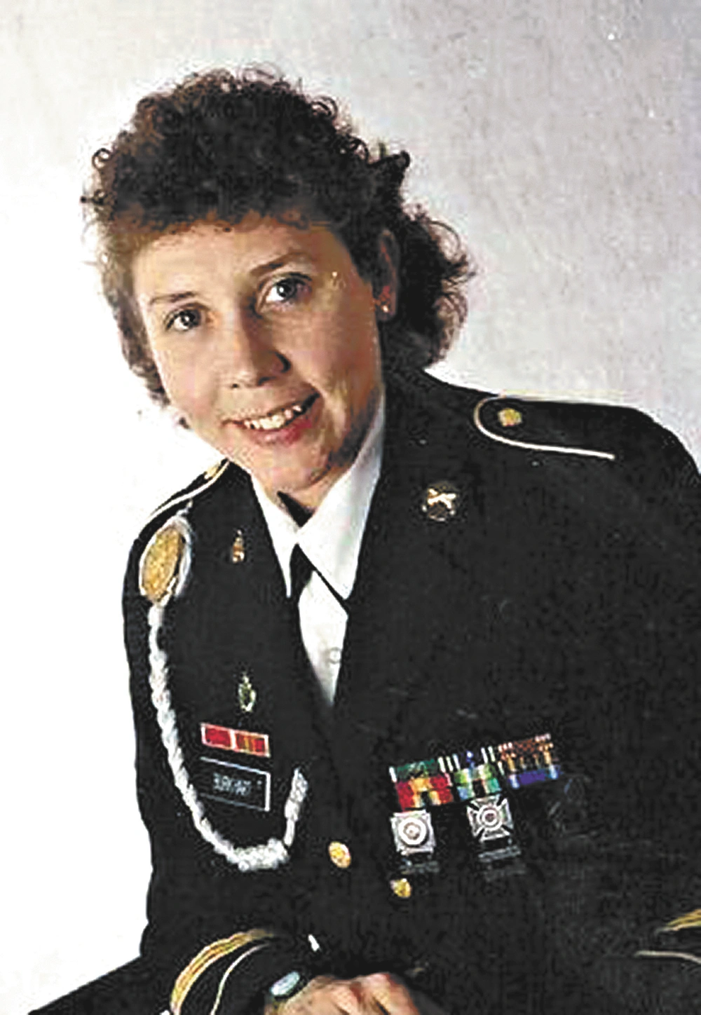 A person in a dark military uniform with insignia, medals, and shoulder patches seated, smiling against a plain light background.