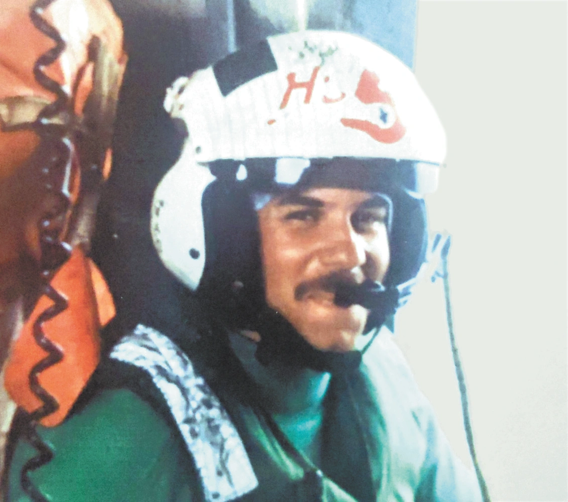 Person wearing white helmet with "H" and red heart, green shirt, vest; another in orange with coiled cord, indoor setting.