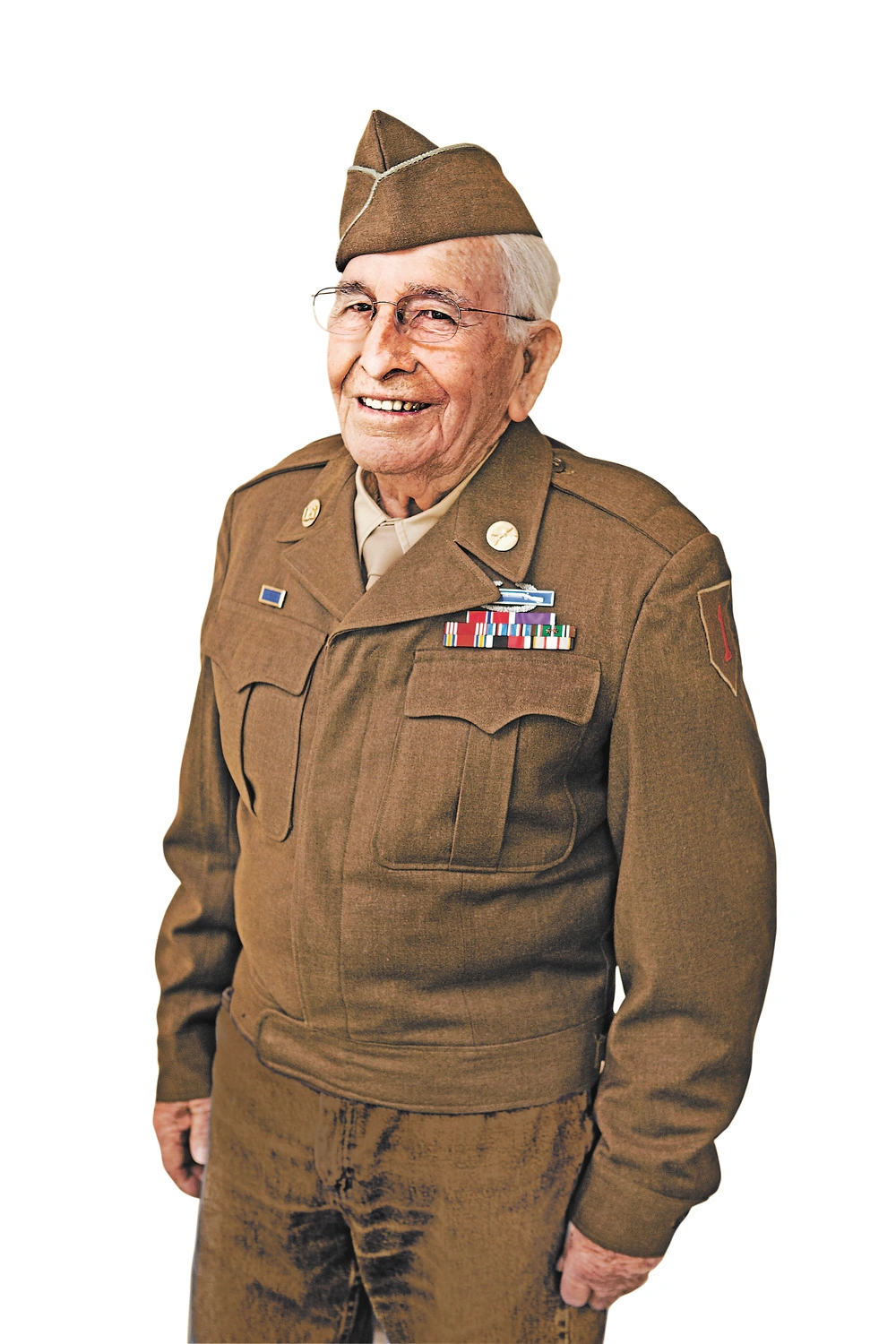 Older man in brown military uniform with cap, medals, and patches stands against plain white background, smiling.